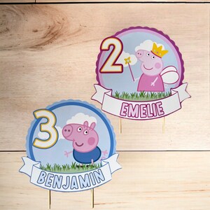 Könnte beinhalten: Zwei Peppa Pig Tortendekorationen, eine blaue und eine rosa. Die blaue Dekoration hat eine Nummer 3 und den Namen "Benjamin" auf einem Banner. Die rosa Dekoration hat eine Nummer 2 und den Namen "Emelie" auf einem Banner.