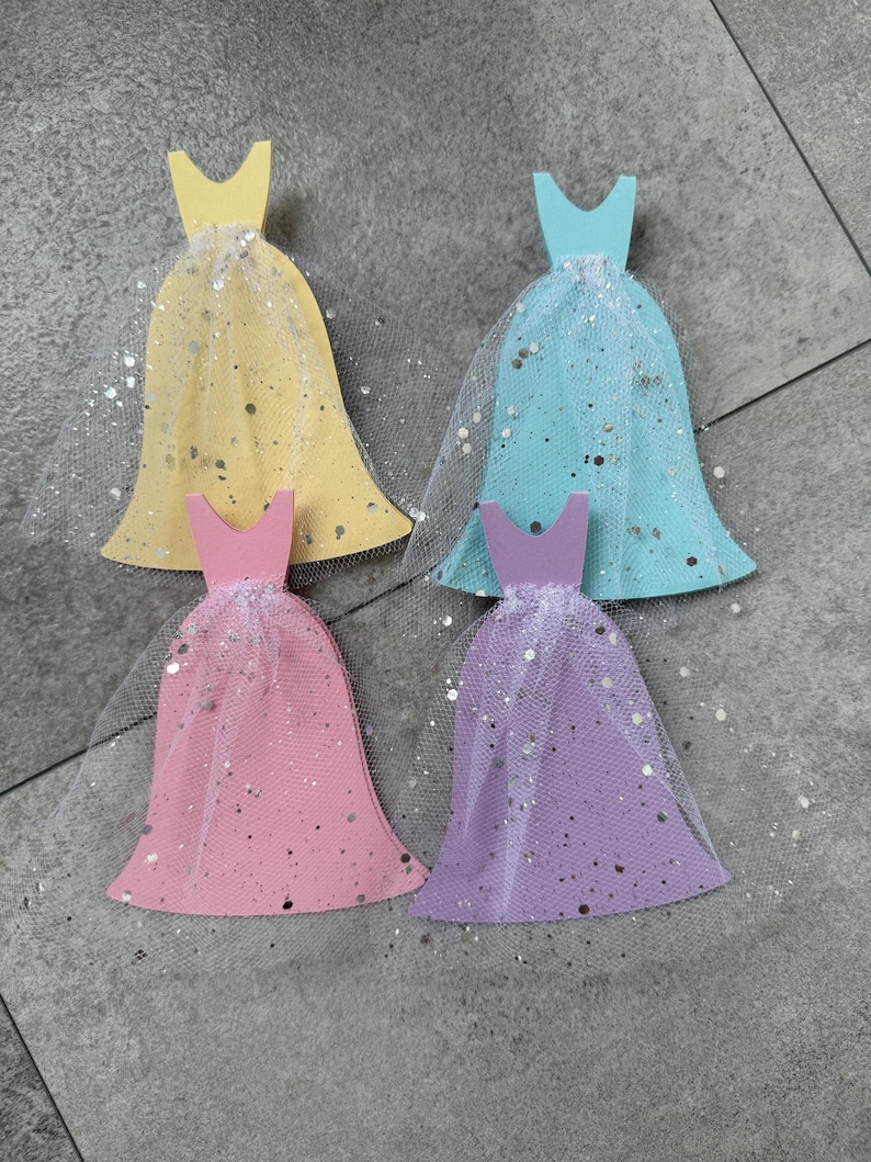 Pu&ograve; includere: Quattro forme di abiti in legno con gonne di tulle e glitter argentati. Gli abiti sono gialli, blu, rosa e viola.