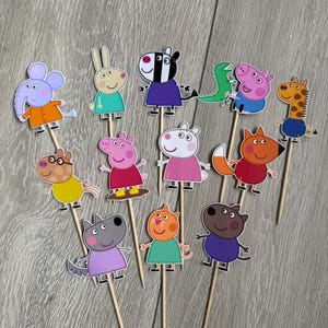 Könnte beinhalten: Eine Sammlung von Cupcake-Toppern mit Cartoon-Charakteren auf Holzstäbchen. Die Topper zeigen verschiedene Tiere, darunter ein Zebra, ein Schwein und eine Giraffe, in leuchtenden Farben. Perfekt zum Dekorieren von Cupcakes oder anderen Backwaren.