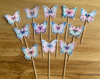 Puede incluir: Un conjunto de adornos para pasteles con forma de mariposa. Cada mariposa presenta un diseño único de acuarela con tonos de rosa, azul y morado. Los adornos están unidos a palitos de madera, perfectos para decorar pasteles o cupcakes.