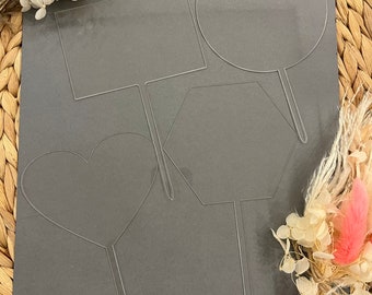 Puede incluir: Cuatro adornos transparentes para tartas de acrílico en varias formas: un rectángulo, un círculo, un hexágono y un corazón. Cada adorno tiene un tallo delgado para insertar en la tarta. Se muestran sobre una superficie gris con flores secas.