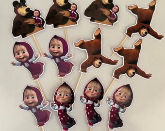 Puede incluir: Adornos para cupcakes con personajes de dibujos animados. Los adornos muestran un oso pardo sosteniendo una muñeca pequeña y una niña con un vestido rojo y un pañuelo en la cabeza. Los adornos están sujetos a palitos de madera.