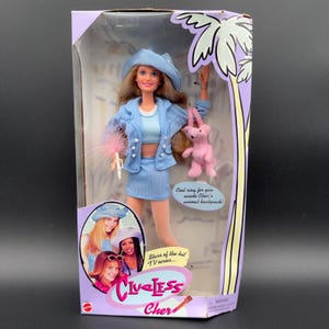 Op de afbeelding: Een "Clueless Cher" pop in de doos. De pop draagt een lichtblauwe outfit en een bijpassende hoed. Ze houdt een roze plumeau en een roze teddybeer-rugzak vast. De doos toont afbeeldingen van de sterren van de tv-serie.