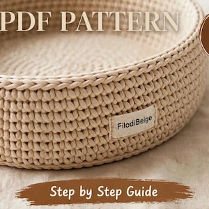 Può includere: Un cesto rotondo all'uncinetto color beige con un motivo a punti visibile e una piccola etichetta in tessuto con la scritta FilodiBeige. L'immagine include il testo PDF PATTERN, Digital Download e Step by Step Guide con un'icona a zampa.