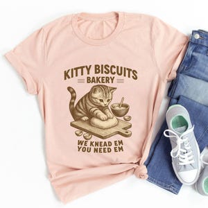 May include: A light pink t-shirt featuring a graphic of a cat kneading dough on a wooden cutting board. The text "Kitty Biscuits Bakery" and "We Knead Em You Need Em" are printed above and below the graphic in brown.