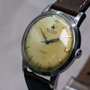 May include: Vintage Buren Grand Prix Power Master watch with a cream-coloured face, silver case, and brown leather strap. The watch face features the brand name and the words "Power Master". The watch has silver hands and hour markers.
