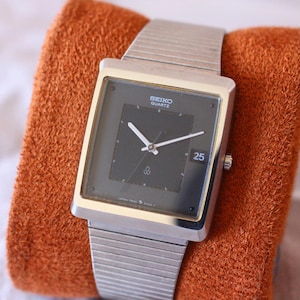 Op de afbeelding: Een zilverkleurig Seiko Quartz horloge met een vierkante zwarte wijzerplaat en een datumweergave met het nummer 25. Het horloge heeft een zilveren band en wordt gepresenteerd op een getextureerd oranje oppervlak.