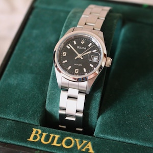Peut inclure: Une montre Bulova à quartz avec un cadran noir, des aiguilles argentées et un guichet de date. Le bracelet est en acier inoxydable et la montre est présentée dans un écrin en velours vert. Le cadran affiche le mot "Bulova".