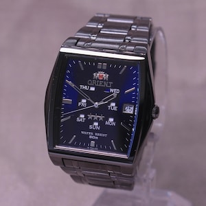 May include: A rectangular Orient watch with a dark blue face and silver hands. The watch displays the day and date, with a silver band. The watch face has the Orient logo and the words "WATER RESIST 50m".