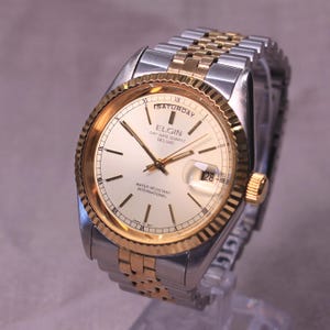May include: A gold and silver Elgin Day-Date Quartz Deluxe watch. The watch face is cream-colored with gold accents, black markers, and the date displayed at the 3 o'clock position. The watch band is a combination of gold and silver links.
