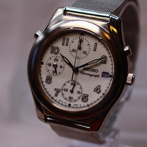 Op de afbeelding: Een goudkleurig Seiko Chronograph horloge met een witte wijzerplaat en zwarte wijzers. Het horloge heeft meerdere sub-wijzerplaten, een datumweergave en een metalen mesh band. De wijzerplaat toont de tekst "SEIKO Chronograph".