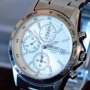 May include: A silver-tone stainless steel Seiko chronograph watch with a white dial, three sub-dials, a date window at the three o'clock position, and a tachymeter scale on the bezel. The watch features luminous hour markers and hands.