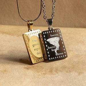 Könnte beinhalten: Zwei rechteckige Anhänger an Ketten. Der goldfarbene Anhänger zeigt den Text "I'm gonna regret this" in einem Oval. Der dunkle Anhänger zeigt eine Tornado-Grafik mit dem mehrfach wiederholten Wort "forever".