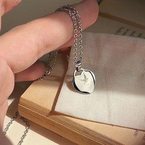 Pu&ograve; includere: Una collana d'argento con un ciondolo a forma di cuore. Il ciondolo presenta un piccolo disegno di un uccello e le parole "Ti amo" incise su di esso.