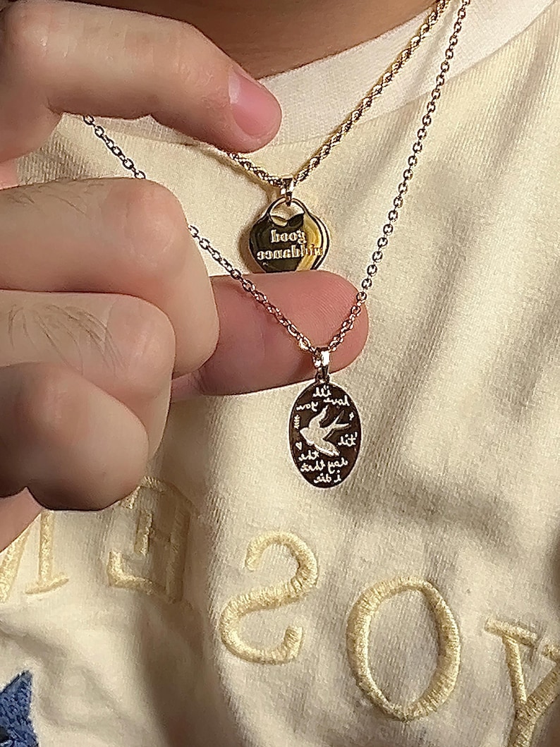 Peut inclure: Deux colliers en or avec des pendentifs. Le collier du haut a un pendentif en forme de c&oelig;ur avec le texte "baby wannabe" inscrit dessus. Le collier du bas a un pendentif rond avec un motif d'oiseau et du texte en arabe.