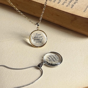 Könnte beinhalten: Zwei Halsketten mit runden Anhängern werden gezeigt. Eine hat eine goldene Kette, die andere eine silberne Kette. Jeder Anhänger trägt den Text "Your delicate point of view" in einem durchsichtigen Glaskreis.