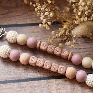 Personalised dummy clips with dusty pink silicone letter beads, wooden beads, and crochet elements, displayed on a wooden surface with dried flowers.