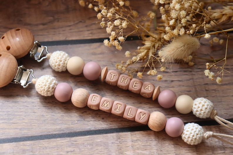 Personalised dummy clips with dusty pink silicone letter beads, wooden beads, and crochet elements, displayed on a wooden surface with dried flowers.