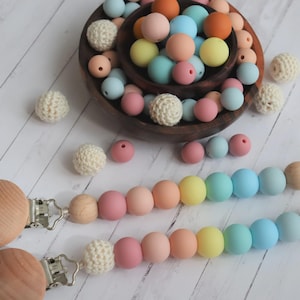 Op de afbeelding: Twee houten fopspeenclips met regenboogkleurige siliconen kralen en haakaccenten. De afbeelding toont ook een houten kom gevuld met pastelkleurige kralen en haakballen, alles op een wit houten oppervlak.