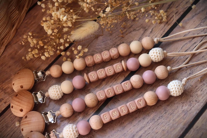 Personalised dummy clips with dusty pink silicone letter beads, wooden beads, and crochet elements, displayed on a wooden surface with dried flowers.