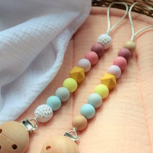 Puede incluir: Dos clips para chupete de bebé con un degradado de cuentas de colores, incluyendo rosa, amarillo y azul, y clips de madera. También se ve un chupete gris. Los clips están sobre una superficie color melocotón.