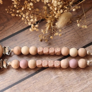 Personalised dummy clips with dusty pink silicone letter beads, wooden beads, and crochet elements, displayed on a wooden surface with dried flowers.
