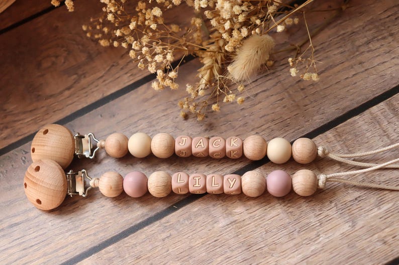 Personalised dummy clips with dusty pink silicone letter beads, wooden beads, and crochet elements, displayed on a wooden surface with dried flowers.