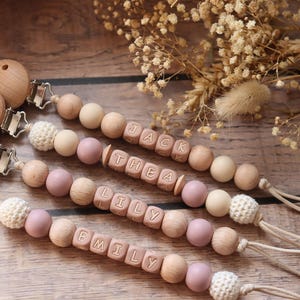 Personalised dummy clips with dusty pink silicone letter beads, wooden beads, and crochet elements, displayed on a wooden surface with dried flowers.