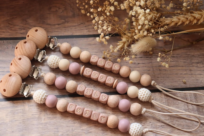 Personalised dummy clips with dusty pink silicone letter beads, wooden beads, and crochet elements, displayed on a wooden surface with dried flowers.
