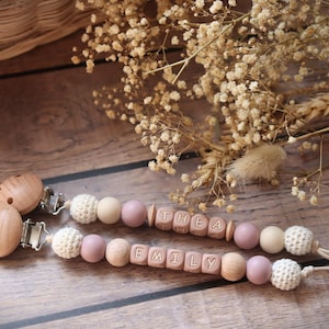 Personalised dummy clips with dusty pink silicone letter beads, wooden beads, and crochet elements, displayed on a wooden surface with dried flowers.