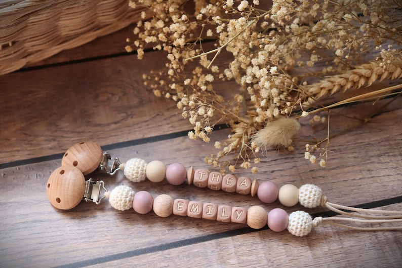 Personalised dummy clips with dusty pink silicone letter beads, wooden beads, and crochet elements, displayed on a wooden surface with dried flowers.