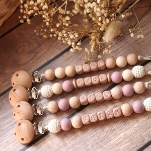 Personalised dummy clips with dusty pink silicone letter beads, wooden beads, and crochet elements, displayed on a wooden surface with dried flowers.
