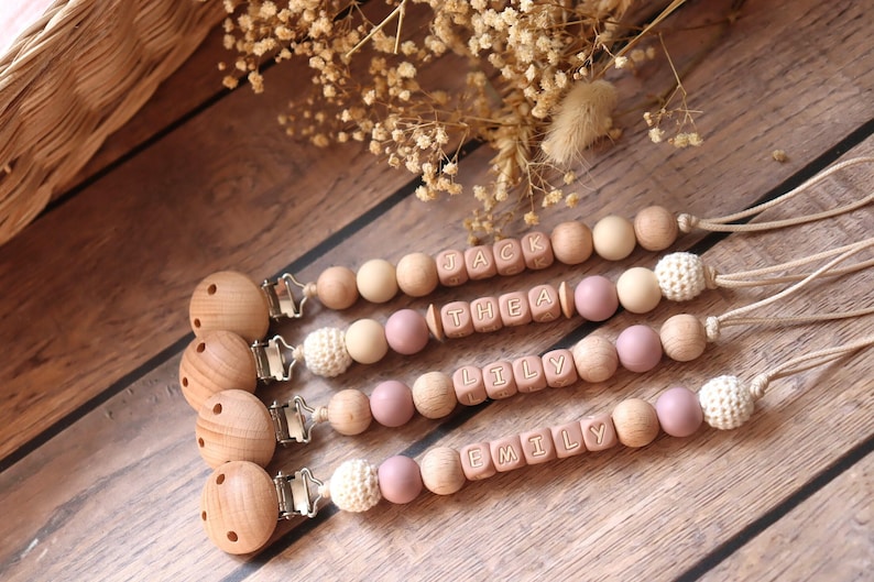 Personalised dummy clips with dusty pink silicone letter beads, wooden beads, and crochet elements, displayed on a wooden surface with dried flowers.