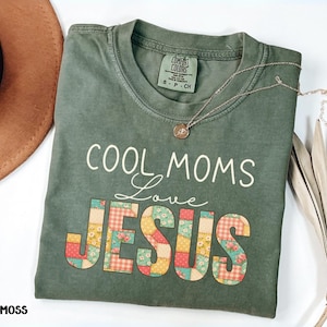 Puede incluir: Una camiseta verde musgo con las palabras "COOL MOMS Love JESUS" en blanco y un diseño de patchwork. Un collar de oro con un colgante descansa sobre la camisa. La camisa está doblada y se muestra con un sombrero marrón y una planta seca.