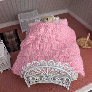 Peut inclure: Scène de chambre de poupée miniature avec un lit en métal blanc et une couverture rose tricotée. Un petit ours en peluche est niché sous la couverture. La chambre comprend une commode blanche, un petit tapis et un panier en osier.
