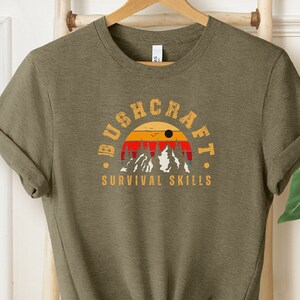 Könnte beinhalten: Ein grünes T-Shirt mit einer Grafik im Vintage-Stil einer Bergkette mit einem Sonnenuntergang dahinter. Der Text "Bushcraft Survival Skills" ist unter der Grafik aufgedruckt.
