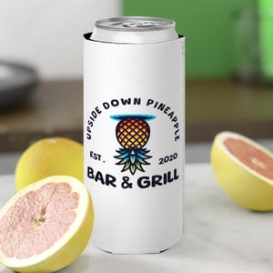 以下が含まれることがあります： 白地に黒と白のロゴが入った缶クーラー。ロゴには「Upside Down Pineapple Est. 2020 Bar & Grill」と書いてあり、青い輪郭のカラフルなパイナップルが描かれている。