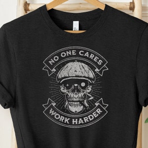 Puede incluir: Camiseta negra con un gráfico blanco de una calavera con una gorra y fumando una pipa. El gráfico incluye el texto "No One Cares Work Harder".