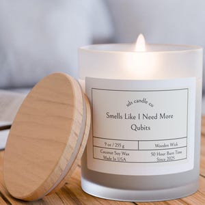 Könnte beinhalten: Ein mattes Glas-Kerzengefäß mit einem Holzdeckel. Die Kerze ist weiß und hat einen Holzdocht. Das Etikett lautet "sds candle co Smells Like I Need More Qubits 9 oz / 255 g Coconut Soy Wax Made In USA Wooden Wick 50 Hour Burn Time Since 2025".
