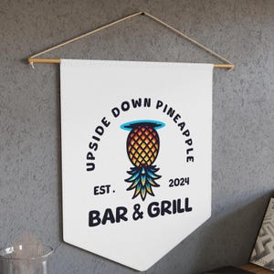 May include: White canvas banner with a black and white logo for a bar and grill called "Upside Down Pineapple Bar & Grill" established in 2024. The logo features a stylized pineapple with a blue halo, yellow and orange fruit, and green leaves.