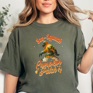 Könnte beinhalten: Ein grünes T-Shirt mit einem Aufdruck eines Sasquatch, der einen Kürbis hält. Der Text "Sas-Squash Pumpkin Patch" ist auf dem T-Shirt aufgedruckt.