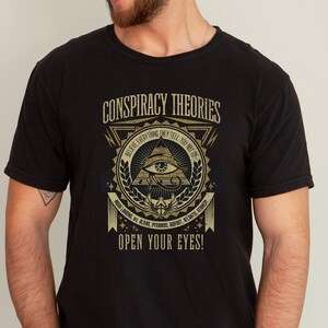 以下が含まれることがあります： 金色のグラフィックデザインが施された黒いTシャツ。ピラミッドの中に、すべてを見通す目が描かれている。デザインの上部には「CONSPIRACY THEORIES」というテキストが書かれている。ピラミッドの周りを「BELIEVE EVERYTHING THEY TELL YOU NOT 100」というテキストが囲んでいる。ピラミッドの下部を「MOON LANDING SIL ALIENS, PYRAMIDS, BIGFOOT, ATLANTIS, KENNEDY」というテキストが囲んでいる。デザインの下部には「OPEN YOUR EYES!」というテキストが書かれている。