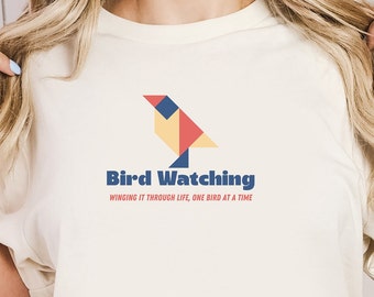 May include: A white t-shirt with a geometric bird design in red, yellow, and blue. The text "Bird Watching" is below the bird, and the text "Winging it through life, one bird at a time" is below that.