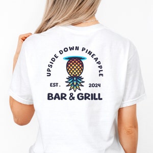 Op de afbeelding: Wit T-shirt met een blauwe, gele en oranje ananas afbeelding. De tekst "Upside Down Pineapple" staat in een cirkel rond de ananas. De tekst "Est. 2024 Bar & Grill" staat onder de ananas.