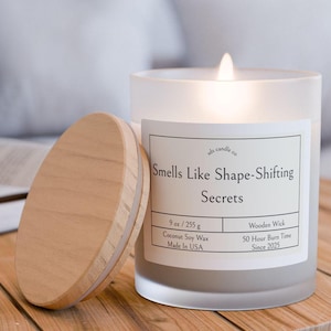 以下が含まれることがあります： 木製の蓋が付いた、フロスト加工されたガラス製のキャンドルジャー。キャンドルには「Smells Like Shape-Shifting Secrets」というラベルが貼られており、「9 oz / 255 g」、「ココナッツソイワックス」、「アメリカ製」、「木製芯」、「50時間燃焼時間」、「2025年から」などの詳細が記載されています。