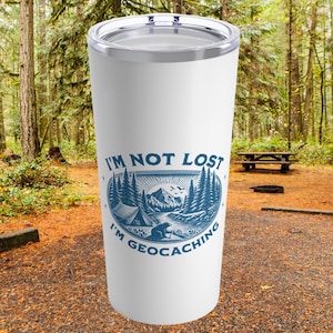 May include: White stainless steel tumbler with a blue and white graphic of a forest scene and the text "I'm not lost I'm geocaching".
