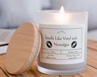 Puede incluir: Un tarro de vela de vidrio esmerilado con una tapa de madera. La vela está encendida y la etiqueta dice "As candle co Smells Like Vinyl and Nostalgia 9 oz/255 g Soy Wax Made In USA Wooden Wick 50 Hour Burn Time Since 2025".