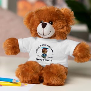 以下が含まれることがあります： カラフルなパイナップルのグラフィックと「Upside Down Pineapple Est. 2020 Swing-N-Stuffy」のテキストが書かれた白いTシャツを着た茶色のテディベア。