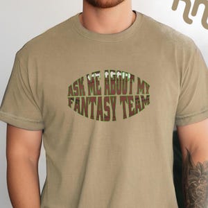May include: A tan t-shirt with the text "Ask Me About My Fantasy Team" printed on the front in a brown and green football shape.