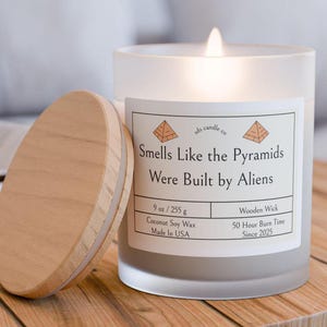 Peut inclure: Un bocal de bougie en verre givré blanc avec un couvercle en bois. La bougie est étiquetée "Smells Like the Pyramids Were Built by Aliens" et possède une mèche en bois. La bougie est faite de cire de soja et de noix de coco et a une durée de combustion de 50 heures. L'étiquette indique également "ads candle co" et "Made in USA".